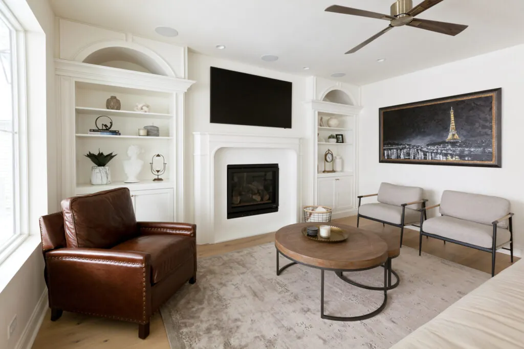 A bright and elegant family room addition featuring built-in white shelving, a central fireplace with a mounted TV above, a brown leather armchair, two modern gray chairs, a round wooden coffee table, and a large Eiffel Tower wall art piece, designed in a clean, contemporary style in the Chicagoland area.