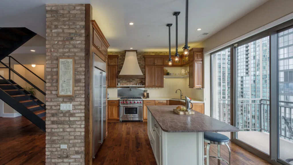Modern kitchen renovation by Design First Builders featuring exposed brick accent wall, custom wood cabinets, granite countertops, and city skyline views in Chicago Illinois