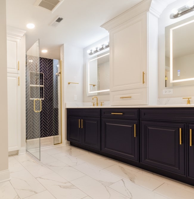 Luxury master bathroom renovation by Design First Builders featuring navy double vanity, gold fixtures, herringbone tile shower, marble flooring, and white upper cabinets in Illinois
