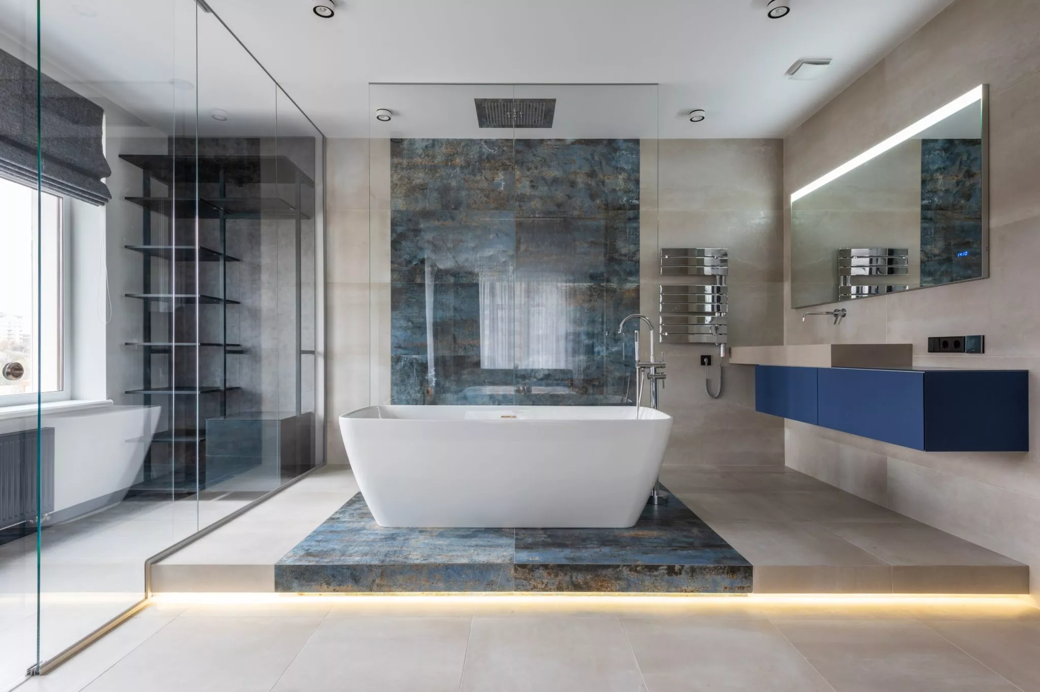 Luxurious modern bathroom featuring white freestanding bathtub on blue stone platform, glass shower enclosure, floating blue vanity, and dramatic stone accent wall
