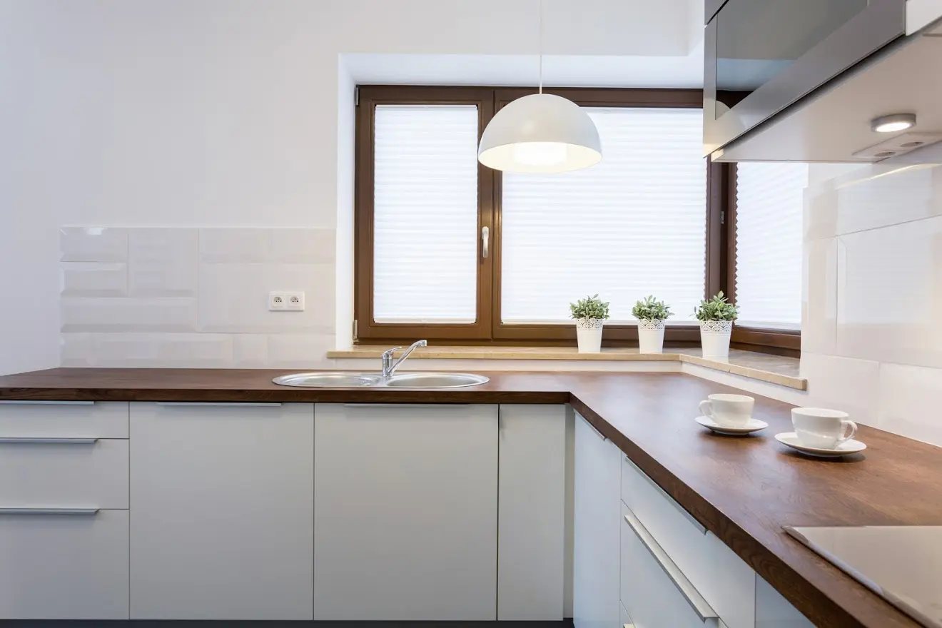 Minimalist white kitchen with walnut wood countertops, corner sink placement, pendant lighting, and wood-framed windows by Design First Builders